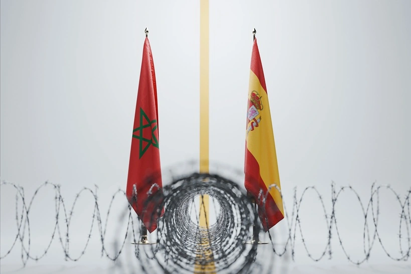 Moroccan and Spanish flags separated by a yellow line behind sharp coiled barbed wire on a white background. Conceptual image of illegal migration crisis, Melilla fence, and border security. Source: Shutterstock