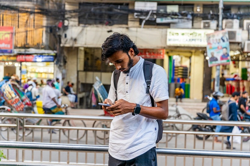 Bangladesh – November 15, 2024: Bengali men browsing, typing phone wearing white half shirt, smart watch in Dhaka road. Source: Shutterstock